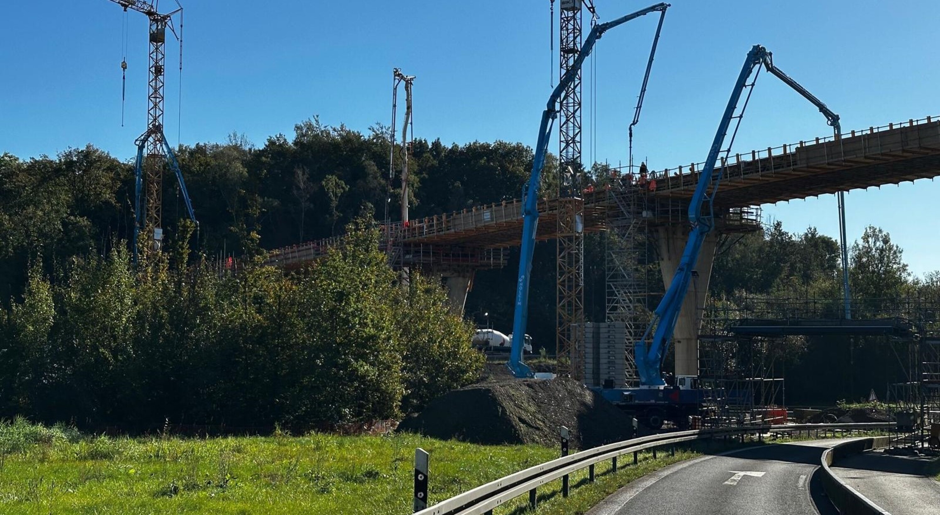 Eine Brückenbaustelle im Grünen: Mehrere hohe Betonpfeiler tragen eine im Bau befindliche Brücke, auf der Bauarbeiter und mehrere große blaue Betonpumpen im Einsatz sind. Im Vordergrund verläuft eine Straße mit Leitplanken und einem Tempolimit-Schild. Die Szene ist von Bäumen und dichtem Grün umgeben, der Himmel ist klar und blau.

                                      
